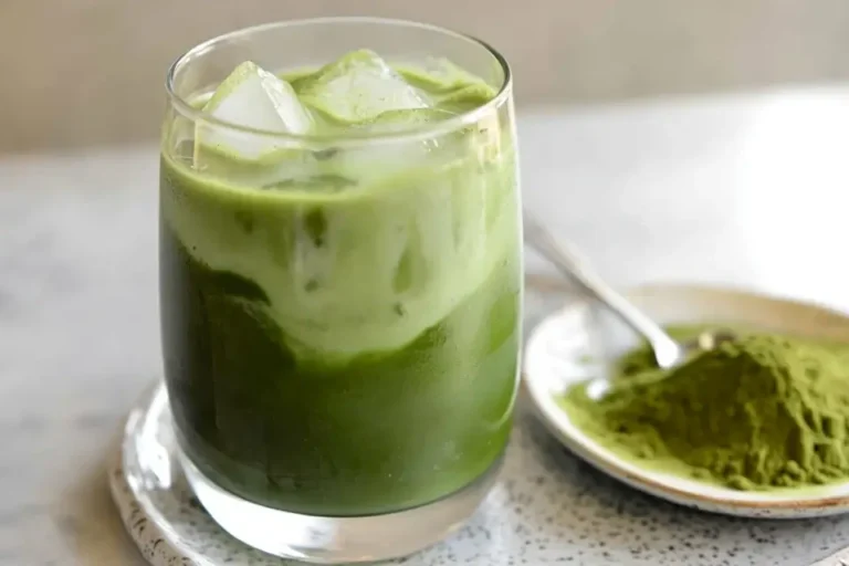 A glass of iced matcha latte with a creamy swirl, next to a plate of matcha powder.